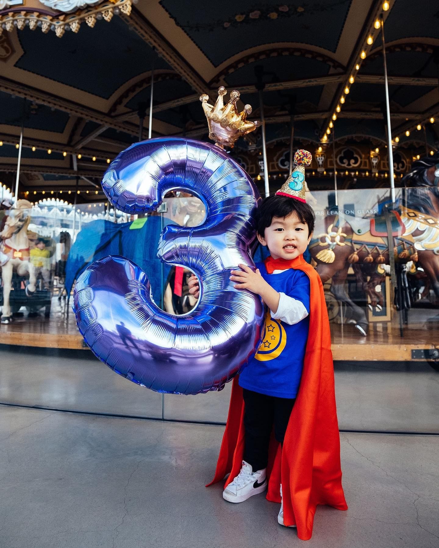 A young child dressed as a superhero holds a shiny, balloon-shaped number three while celebrating their birthday at a carousel, captured by Stylish & Hip Kids Photography. Featured in "10+ Kids Birthday Photographers in New York City."