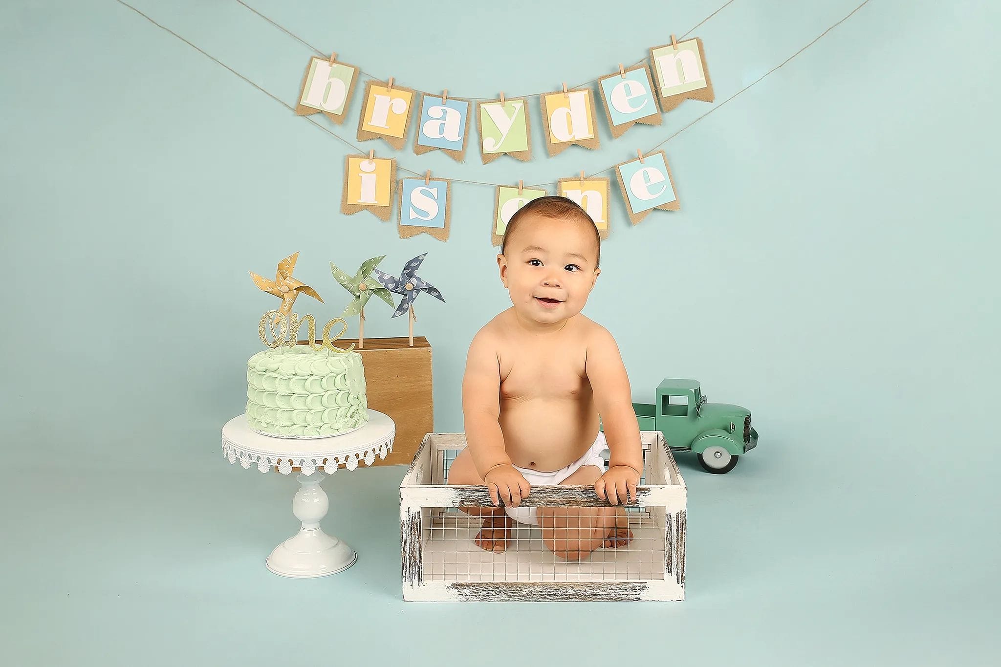 A baby celebrating their first birthday poses in a vintage-inspired setup with a pastel cake, decorative pinwheels, and a banner reading "Brayden is one," captured by Kyos Photography.
