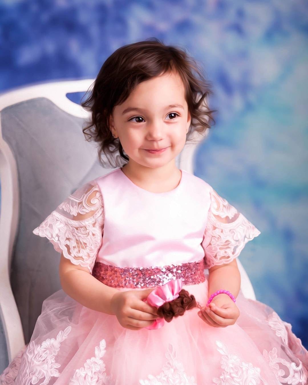 A young girl dressed in an elegant pink gown with lace details sits on a chair, smiling softly against a dreamy blue backdrop, captured by Julie Pavlova Photography.