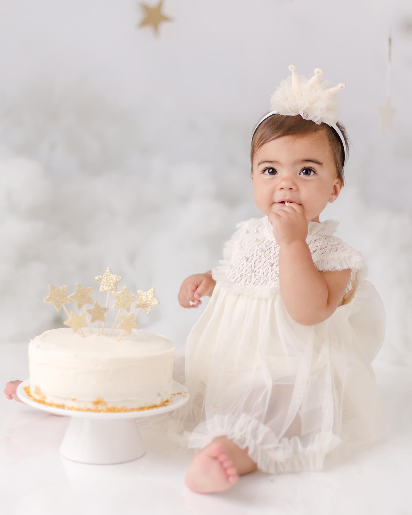 A baby dressed in a delicate white outfit with a crown headband sits beside a white cake adorned with golden star toppers, playfully tasting the frosting in a dreamy, cloud-like setting, captured by Little New York Photography.