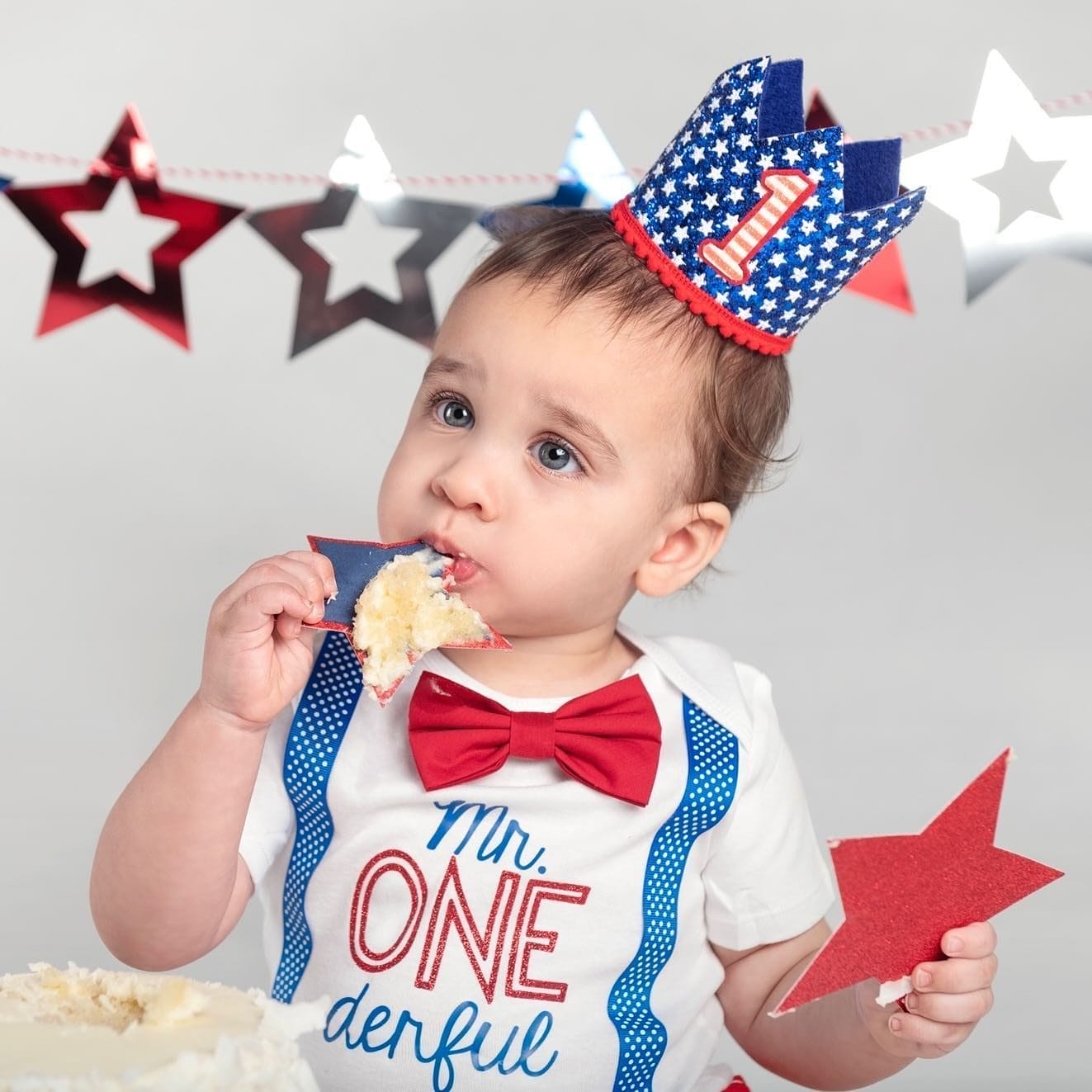 A baby celebrating their first birthday wears a patriotic-themed outfit with a "Mr. ONEderful" shirt, red bow tie, and a star-patterned crown while enjoying a cake smash with red, white, and blue decorations in the background, captured by Classic Kids Photography NYC.