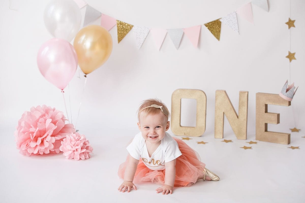A smiling baby dressed in a peach tutu and a "One" shirt crawls in a beautifully decorated birthday setup featuring gold and pink balloons, a festive banner, and large gold letters spelling "ONE," captured by Celimageskids.