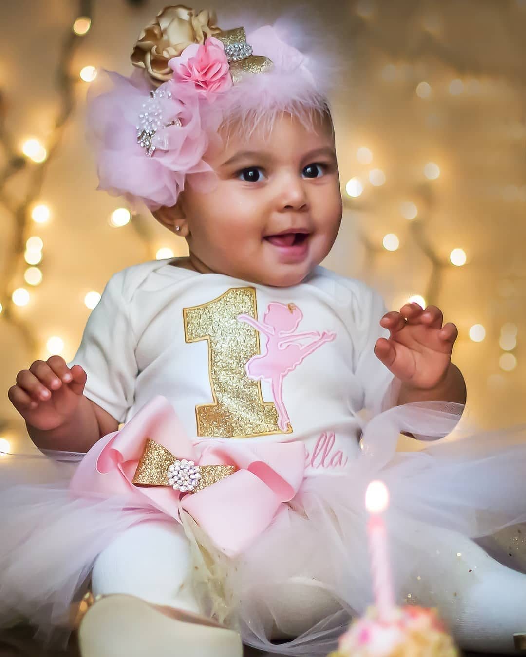 A baby celebrating their first birthday wears a tutu, a festive "1" outfit with a ballerina design, and a decorative floral headpiece, smiling in front of warm twinkling lights, captured by FJA Photography.