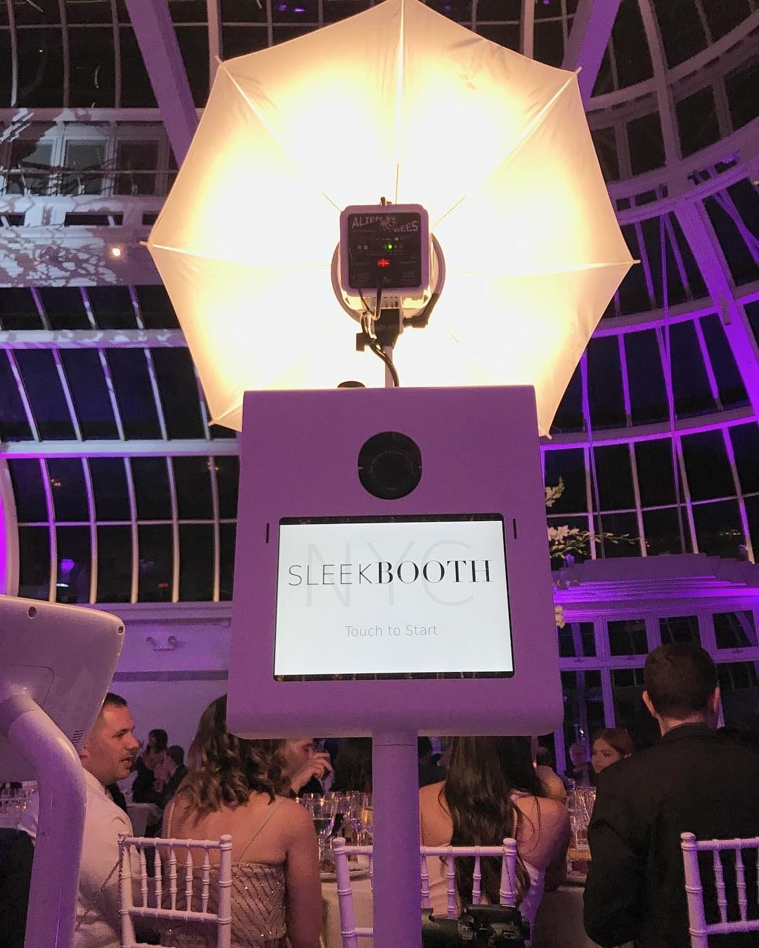 A sleek, modern photo booth setup by Sleek Booth NYC, illuminated by a large umbrella light in a stylish event space. The touchscreen display reads ‘Sleek Booth,’ with guests seated in the background under a glass-domed ceiling. This photo booth is featured in the 15+ Best Photo Booth Rentals in New York City blog post.