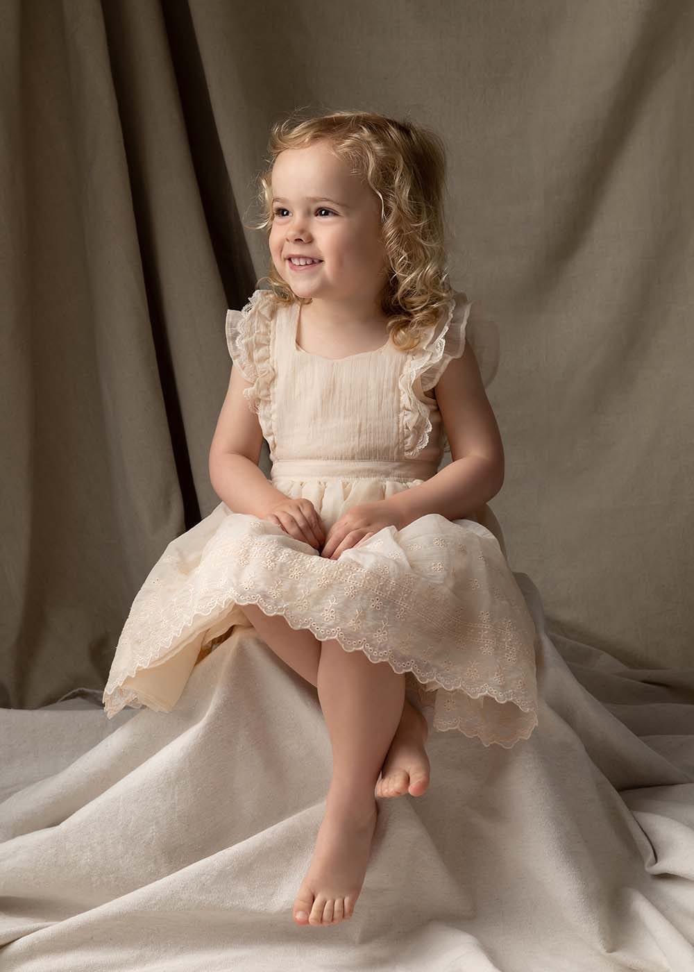 A young girl with curly blonde hair sits gracefully in a soft, neutral-toned setting, wearing a delicate cream-colored dress with lace details, captured by Glow Portraits. Featured in "10+ Kids Birthday Photographers in New York City."