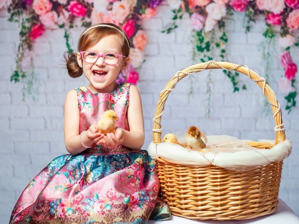 A young girl wearing pink glasses and a colorful floral dress smiles joyfully while holding a baby chick, with a wicker basket of more chicks beside her, set against a bright floral backdrop, captured by PhotoOp NYC.
