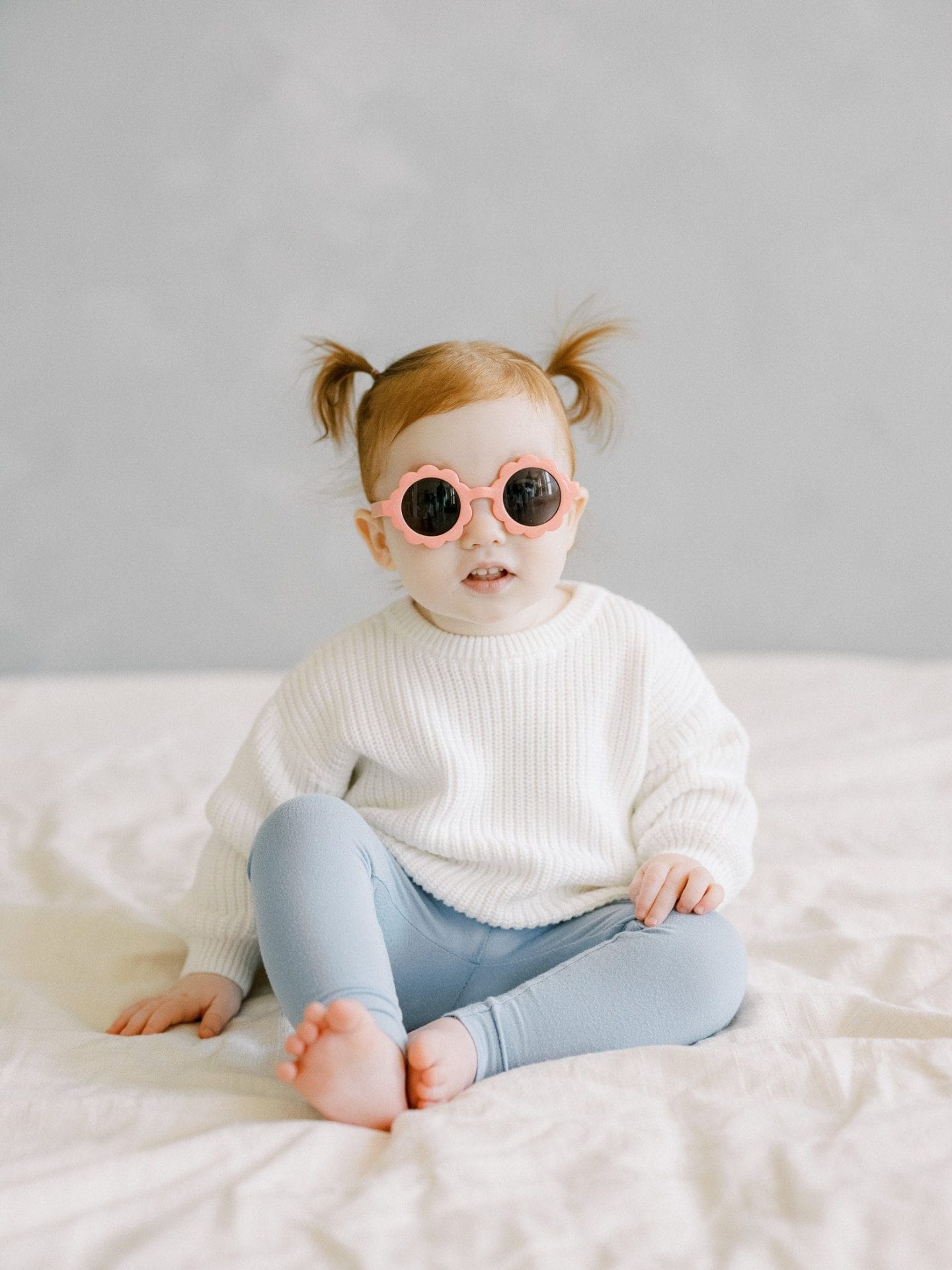 A toddler with pigtails sits on a bed wearing a cozy white sweater, light blue leggings, and playful pink flower-shaped sunglasses, exuding a cool and carefree vibe, captured by Michelle Lange Photography.