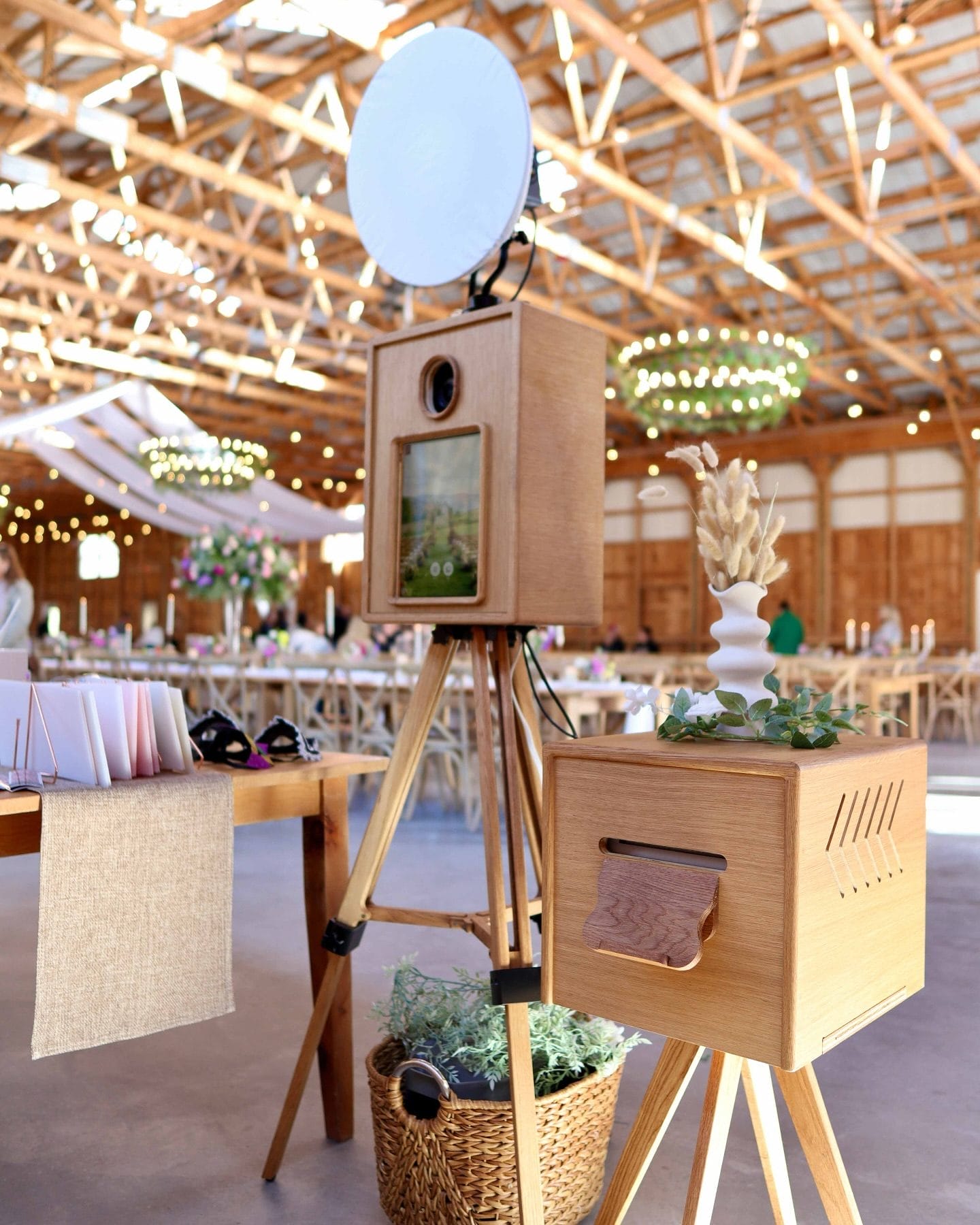 A vintage-style wooden photo booth on a tripod stand from Cinematic Photo Booths, set in a rustic barn venue with string lights and floral accents. The booth features a circular light on top and a separate wooden storage box, creating a warm, timeless aesthetic. Sleek Booth is also highlighted in the 15+ Best Photo Booth Rentals in New York City blog post.