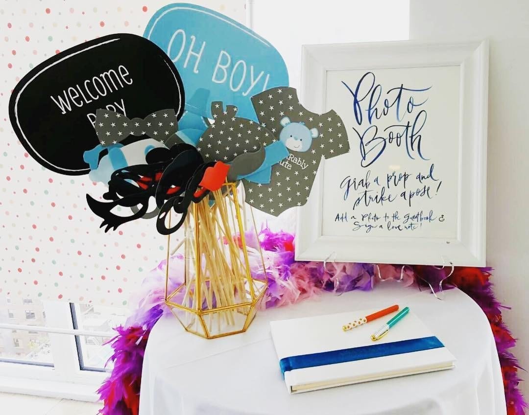 A baby shower photo booth setup by Sweet Booths, featuring a gold geometric holder filled with baby-themed props like speech bubbles. A framed sign encourages guests to ‘grab a prop and strike a pose,’ while a guestbook and pens are placed nearby. This photo booth is highlighted in the 15+ Best Photo Booth Rentals in New York City blog post.